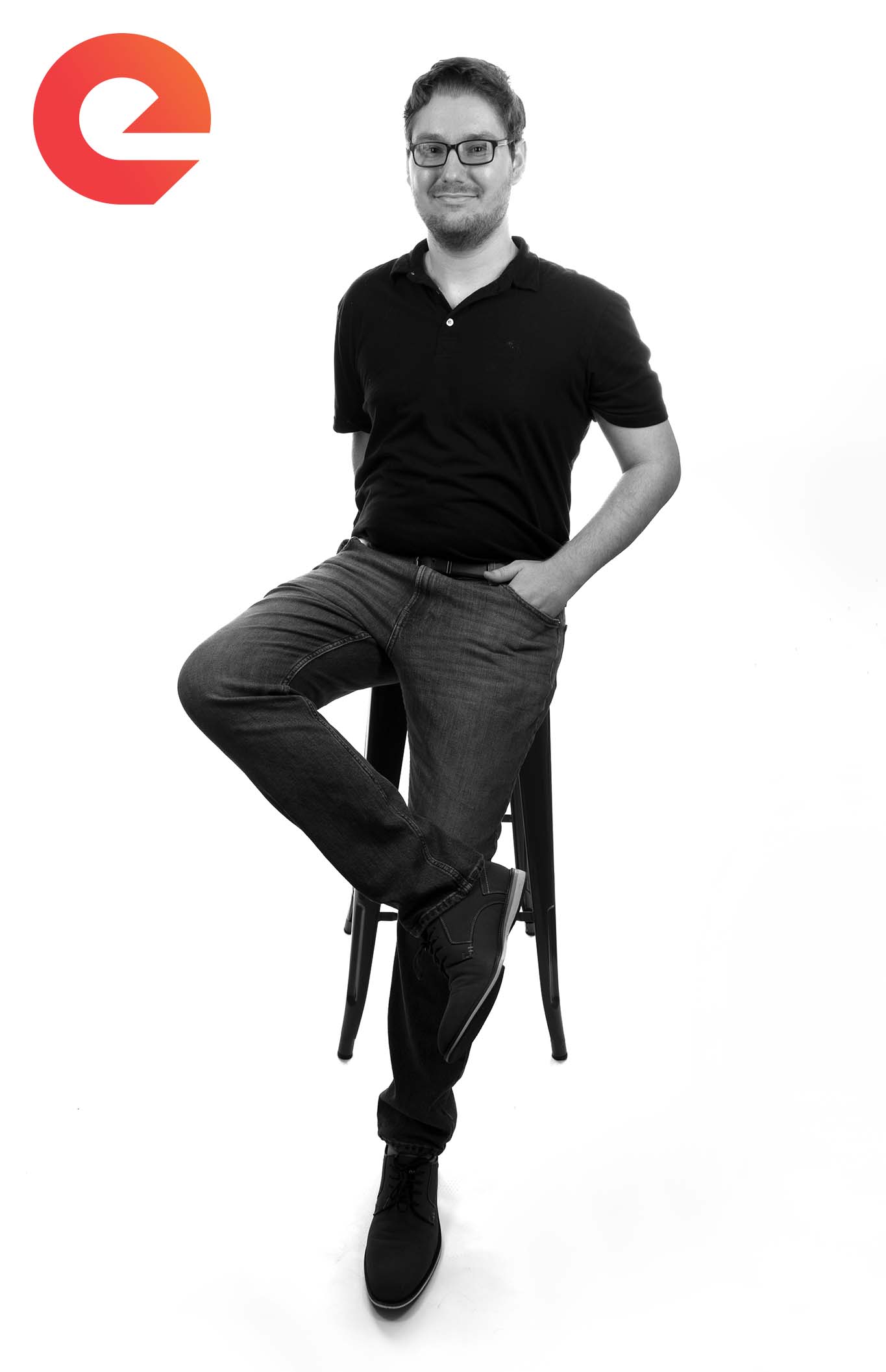 Josh Loufek | Web developer. Black and white photo. White male with black rimmed glasses, shorter tousled dark brown hair, slight facial hair, black polo, dark jeans, black shoes. Leaning casually against a black stool, right leg slightly crossed with the ankle resting on the left knee. Left hand in the pocket. White background.